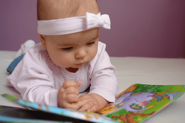 девочка 9 месяцев лежит на животе полу, и внимательно рассматривает книжку