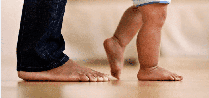 rebenok-shagaet, первые шаги ребенка. на фото - босая нога отца и ножки ребенка - одна стоит на полу, другая опирается на пол большим пальцем ноги