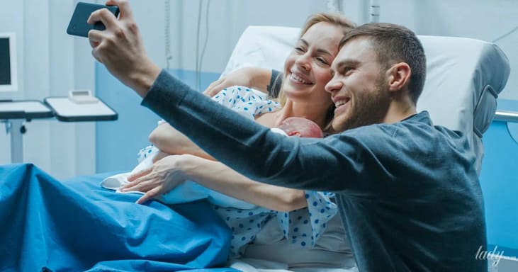 молодой отец фотографирует жену с новорожденным в палате после родов