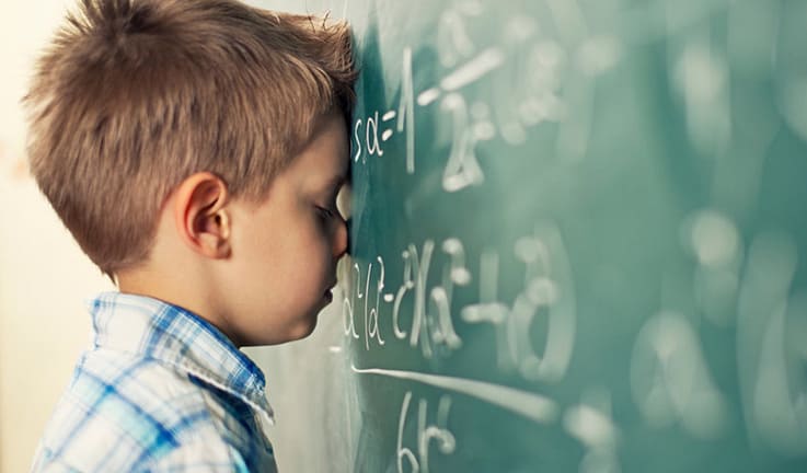 ребенок, страдающий дискалькулией стоит у школьной доски, уперся в нее головой и плачет. На доске написаны математические примеры