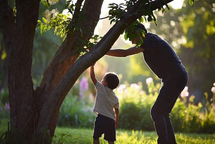 отец и маленький сын стоят у дерева, мальчик хочет на него залезть