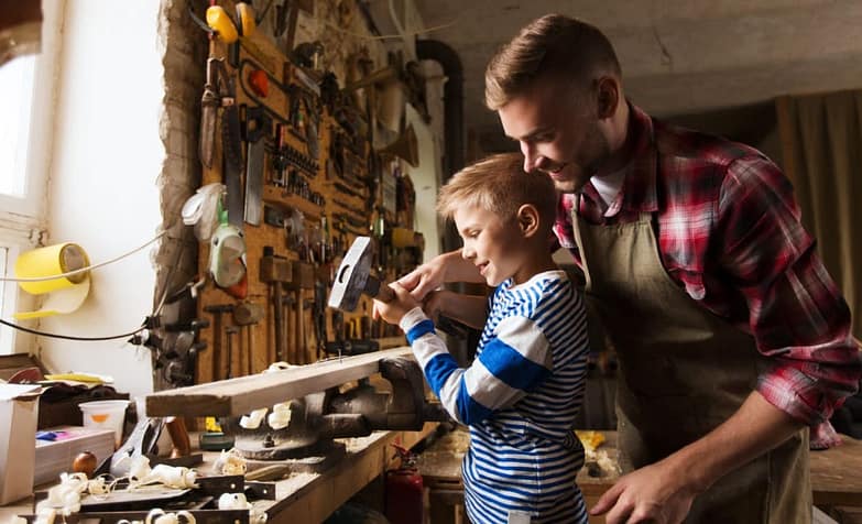мастерская, в ней множество разных инструментов. Отец помогает сыну 10 лет забивать гвоздь в доску молотком