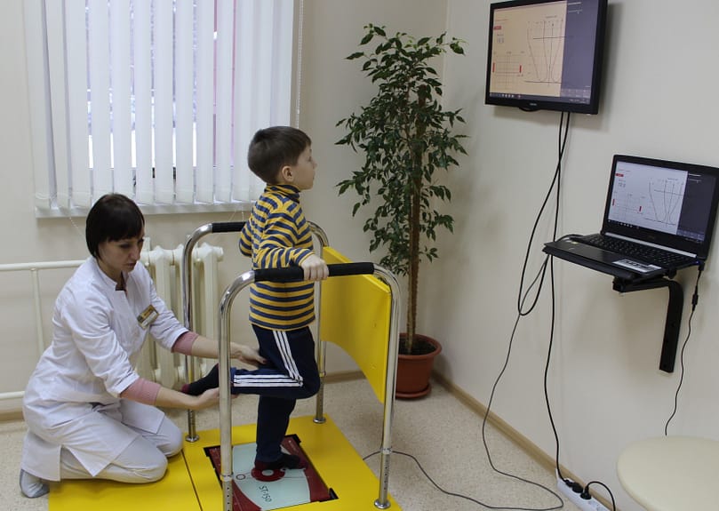 ребенок с диспраксией стоит на стабилоплатформе, врач помогает ему