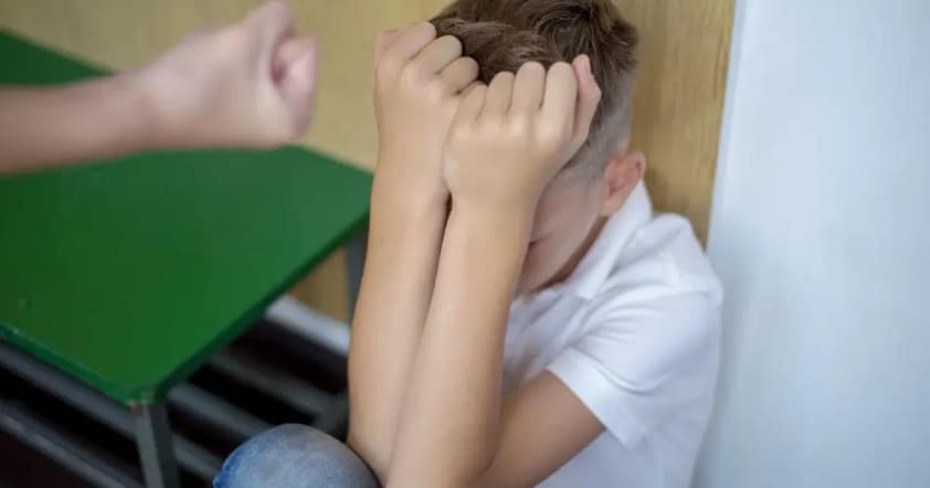 мальчик сидит у стены, закрыл руками лицо, одноклассник пытается его ударить